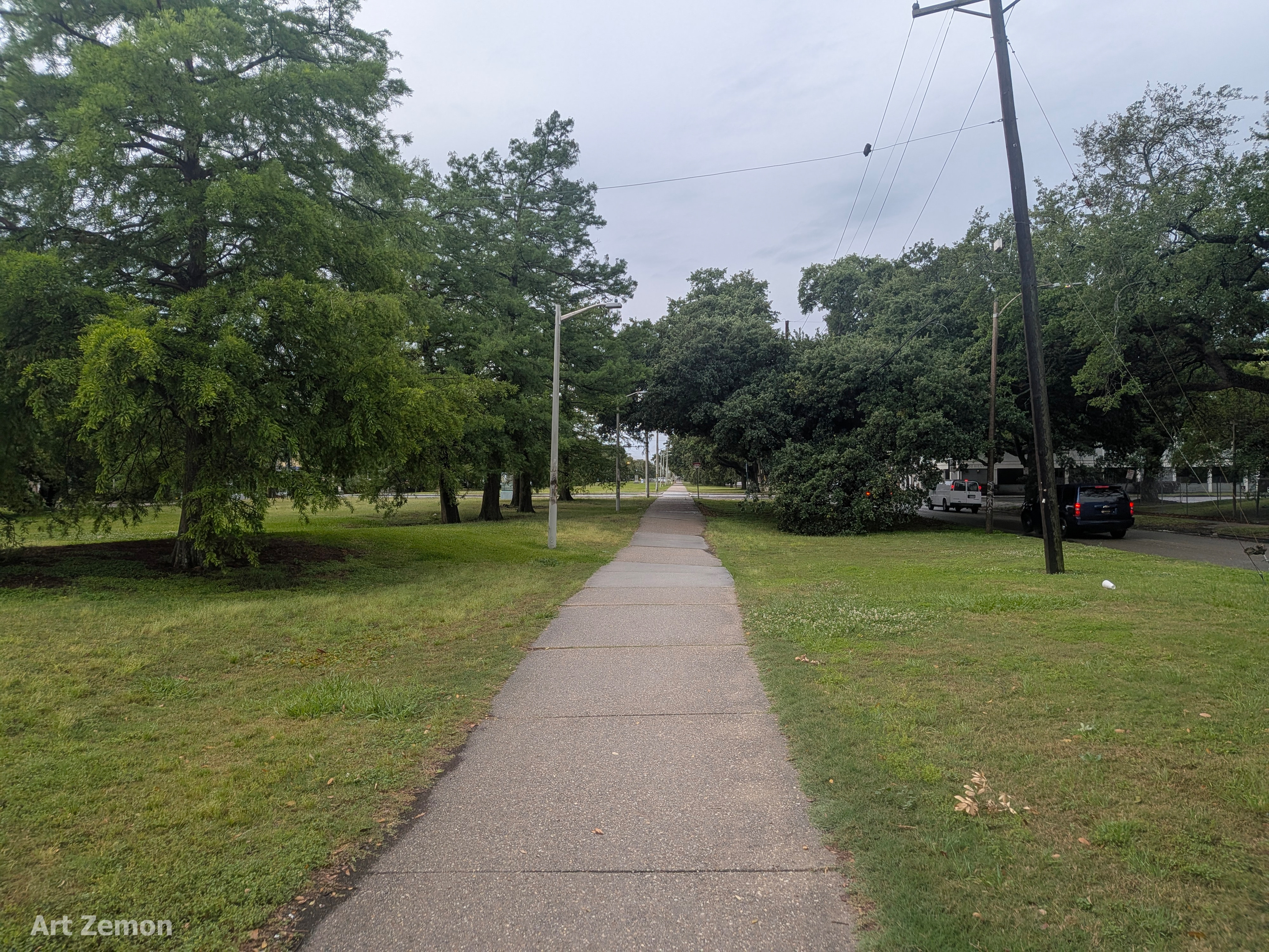 walking/bicycling paved path running from the lower center of the screen directly away into the distance. Grass and trees line the path. There is a street with cars parked at the far right