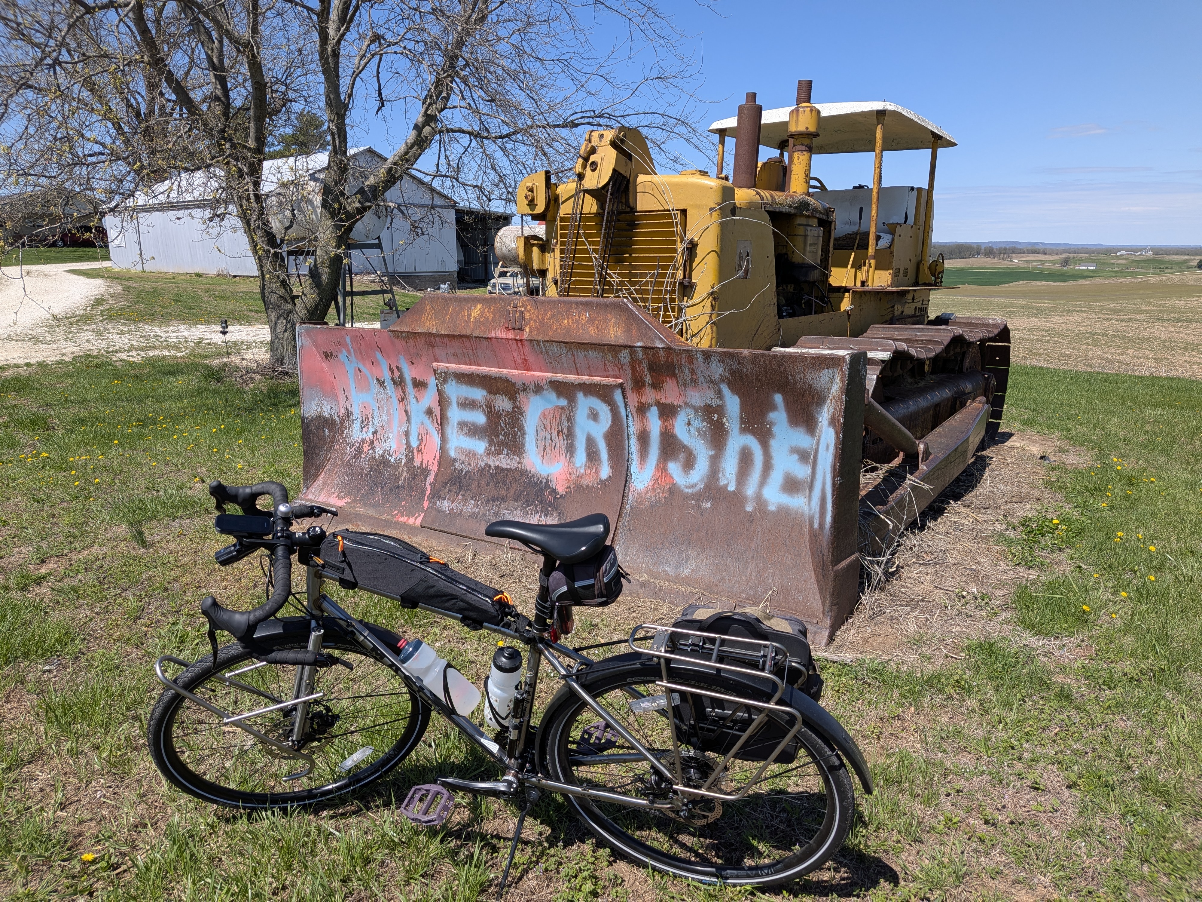 Old bulldozer with "Bike Crusher" painted on the rusted blade. A touring bicycle is parked in front of it, posing for the photo