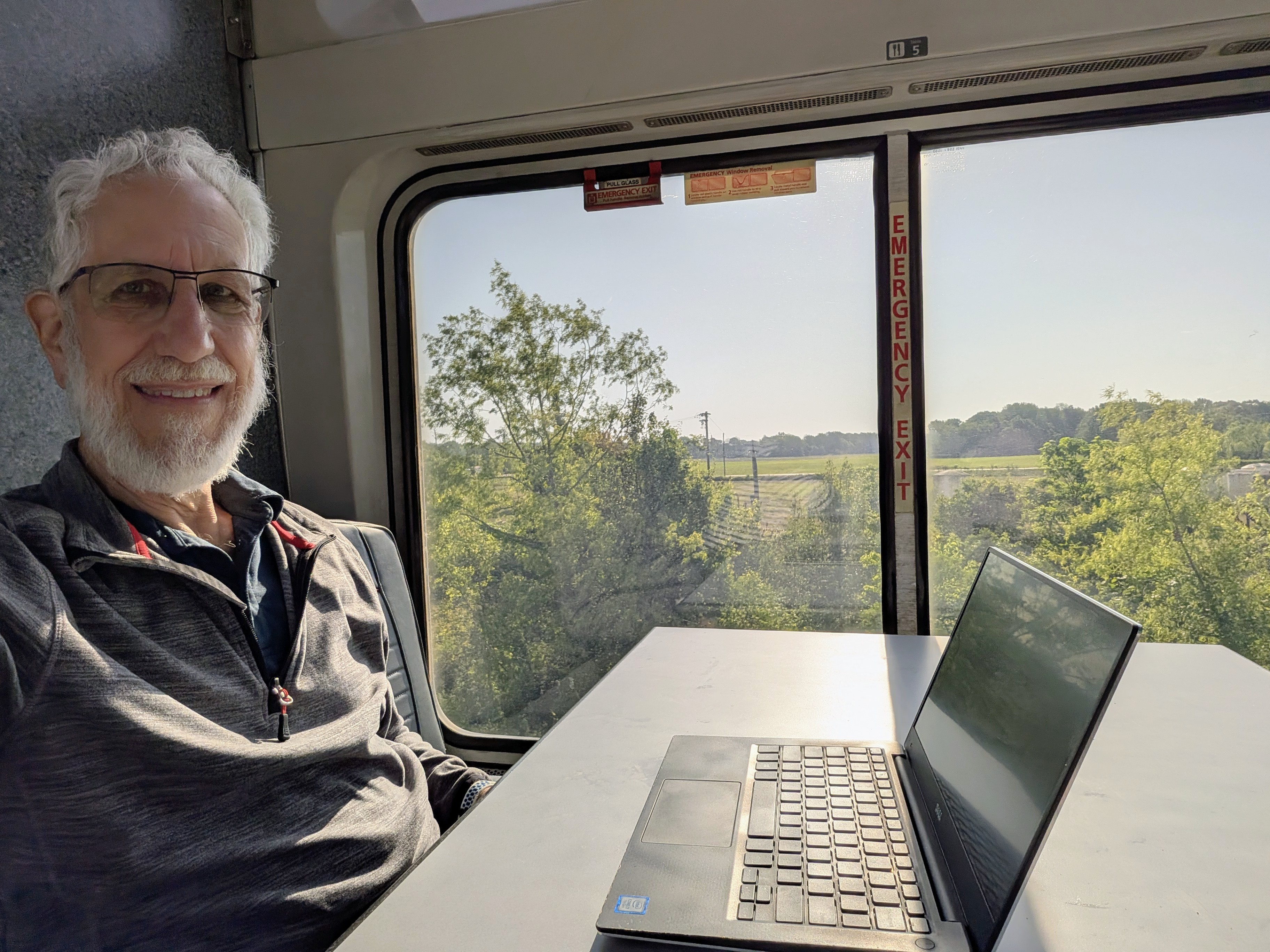 Art Zemon at a table in the observation car of a train. There is a laptop on the table and green fields outside the window